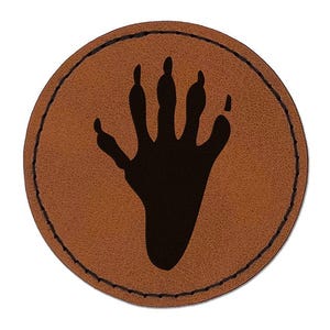 May include: A circular brown patch featuring a black silhouette of a five-fingered animal paw print in the center. The patch has a dark stitched border running along the outer edge, creating a defined circular shape with a textured surface.