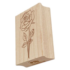 May include: A wooden stamp featuring a detailed outline of a rose with a stem, leaves, and a bud. The stamp is made of light-colored wood and has a curved handle for easy gripping. The rose design is etched in brown.