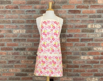 May include: A white apron with a pink and yellow floral pattern. The apron has a single pocket on the front.