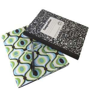 May include: A black and white composition notebook with a green and blue patterned fabric cover. The notebook is on top of the fabric cover and a black pen is lying on top of the fabric cover.