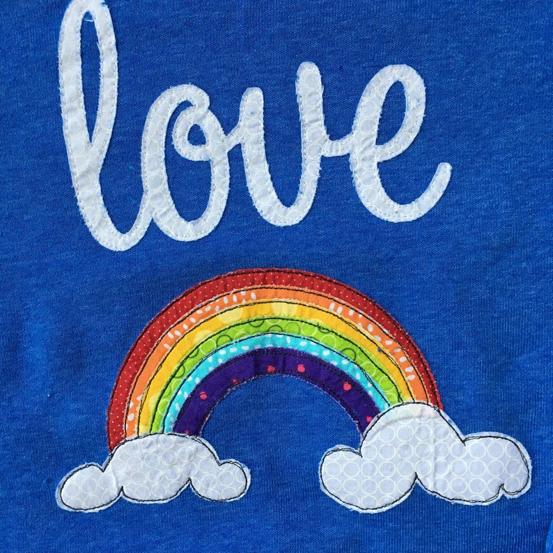 May include: A blue fabric with the word "love" embroidered in white and a rainbow with clouds embroidered in colorful fabrics.