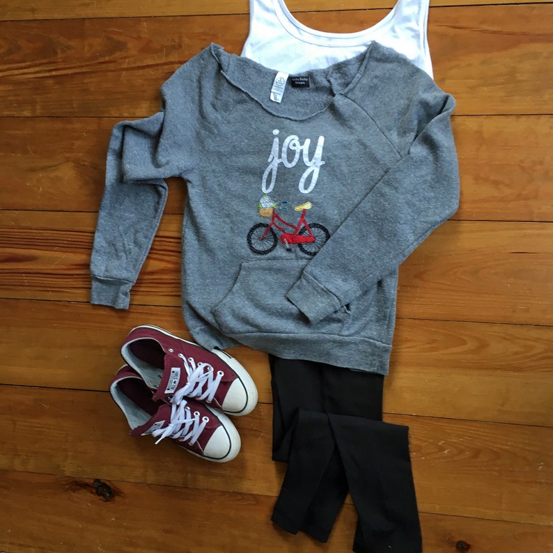 May include: Gray sweatshirt with the word "joy" and a red bicycle with a basket of flowers embroidered on the front. The sweatshirt is paired with black leggings and maroon Converse sneakers.