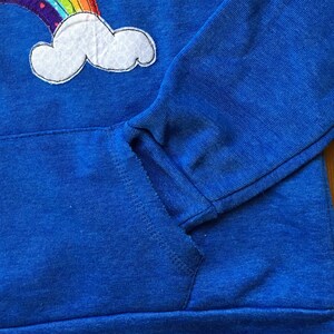 May include: A blue hoodie with a rainbow and cloud applique. The hoodie has a pocket on the front.