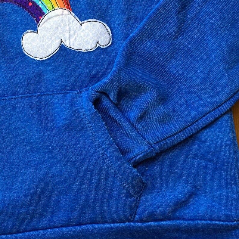 May include: A blue hoodie with a rainbow and cloud applique. The hoodie has a pocket on the front.