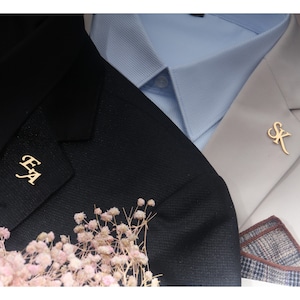 May include: Two gold-toned lapel pins with initials. The pin on the left says "EA" and the pin on the right says "SK".