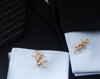 May include: A pair of gold-toned metal cufflinks featuring the letters JFK in a stylized script font. The cufflinks are attached to the light blue, diagonal-textured cuffs of a dress shirt, which is worn under a black pinstriped suit jacket.