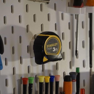 Puede incluir: Un tablero de clavijas blanco con una cinta métrica amarilla y negra colgada. La cinta métrica está etiquetada como "STANLEY TYLON 3m". También hay varios destornilladores y otras herramientas colgadas en el tablero de clavijas.