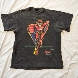 Puede incluir: Una camiseta negra de manga corta con un gráfico del personaje de cómic The Flash corriendo hacia adelante. El personaje viste un traje rojo con un emblema de rayo amarillo. El texto The Flash está impreso en rojo en la parte inferior.