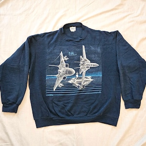 May include: A dark navy blue crewneck sweatshirt featuring a white and light blue graphic print of two F-14 Tomcat fighter jets flying over an aircraft carrier. The text F-14 TOMCAT is printed in white above the jets on the front of the garment.