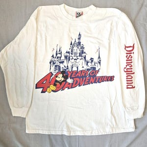 May include: A white long-sleeve t-shirt featuring a graphic of a castle, Mickey Mouse, and the text 40 YEARS OF ADVENTURES on the front. The word Disneyland is printed in red vertically along the left sleeve. The shirt has a crew neckline.