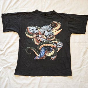 Peut inclure: Un t-shirt noir à manches courtes avec une impression graphique représentant une créature musclée et anthropomorphe ressemblant à un taureau, enlacée par les anneaux d'un grand serpent sombre. La créature a la peau bleue et un museau blanc.