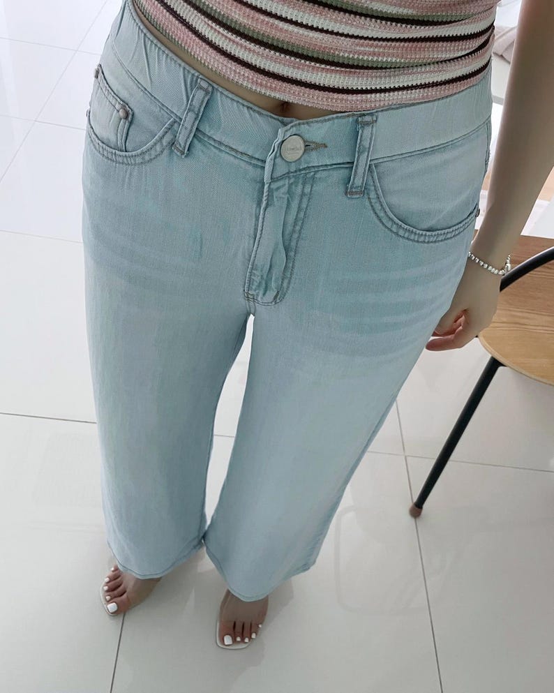 Puede incluir: Vaqueros de mezclilla azul claro con dise&ntilde;o de cintura alta, cierre de bot&oacute;n, cremallera y estilo cl&aacute;sico de cinco bolsillos. Los pantalones tienen un corte recto y un acabado de lavado claro, combinados con un top de punto a rayas.