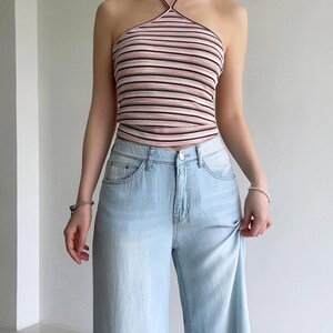 Puede incluir: Un top halter acanalado con rayas horizontales en tonos rosa, blanco y negro, combinado con pantalones vaqueros de mezclilla azul claro. El top tiene un cuello alto con tirantes finos alrededor del cuello y el pantal&oacute;n tiene cierre de bot&oacute;n.