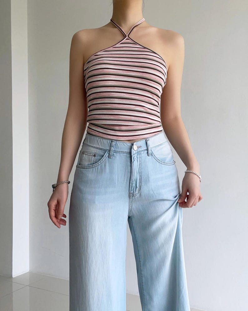 Puede incluir: Un top halter acanalado con rayas horizontales en tonos rosa, blanco y negro, combinado con pantalones vaqueros de mezclilla azul claro. El top tiene un cuello alto con tirantes finos alrededor del cuello y el pantal&oacute;n tiene cierre de bot&oacute;n.