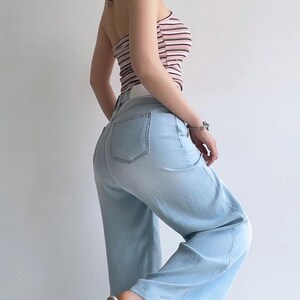 Puede incluir: Un top tipo halter con rayas horizontales en tonos rosa, blanco y negro, combinado con pantalones vaqueros de mezclilla azul claro. Los pantalones tienen un corte de cintura alta, piernas anchas y bolsillos traseros visibles. El conjunto se completa con zapatos de tac&oacute;n bloque transparente.