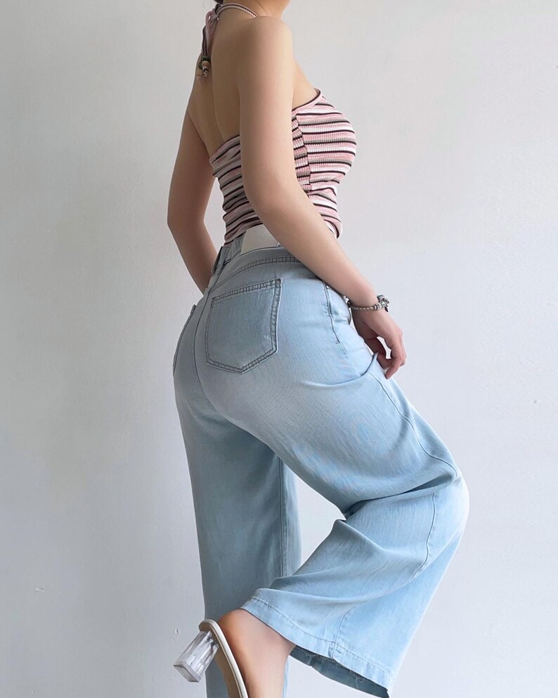 Puede incluir: Un top tipo halter con rayas horizontales en tonos rosa, blanco y negro, combinado con pantalones vaqueros de mezclilla azul claro. Los pantalones tienen un corte de cintura alta, piernas anchas y bolsillos traseros visibles. El conjunto se completa con zapatos de tac&oacute;n bloque transparente.