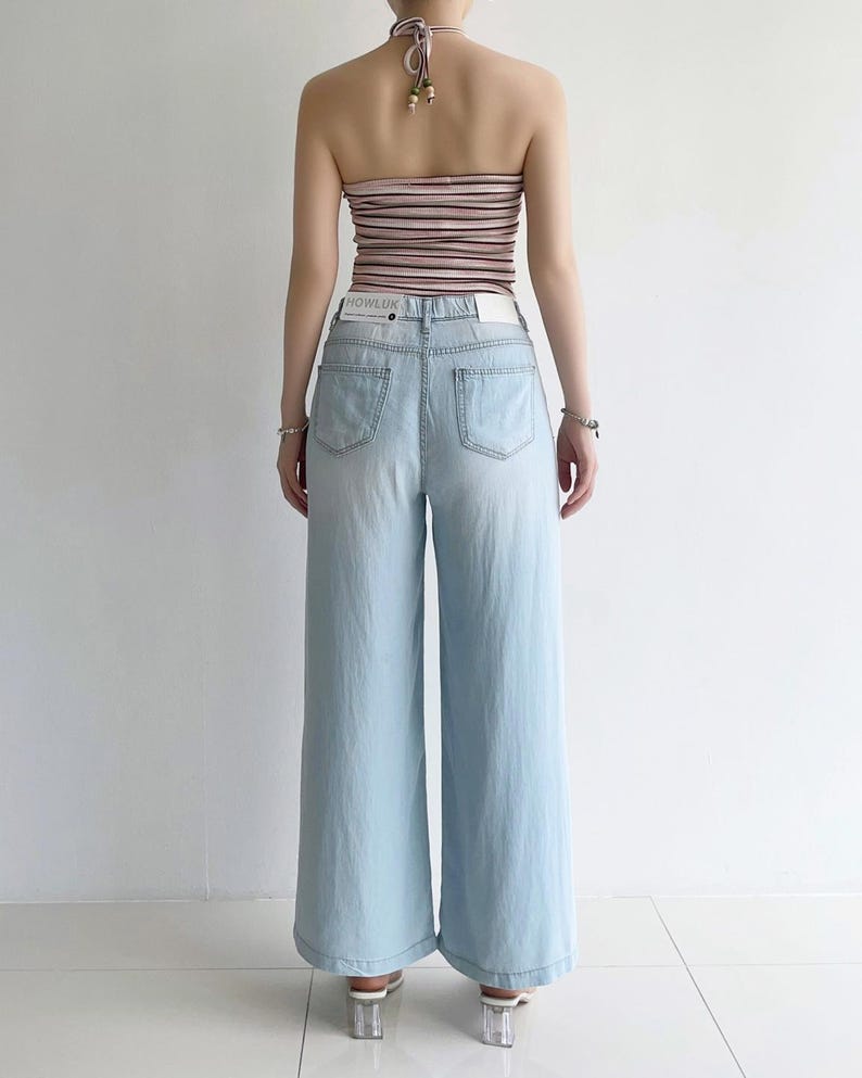 Puede incluir: Unos vaqueros de pernera ancha en color azul claro con cintura alta y dos bolsillos traseros. La cintura lleva una etiqueta blanca con el texto HOWLUK. Los vaqueros se combinan con un top halter de rayas horizontales en tonos rosa, marr&oacute;n y blanco.