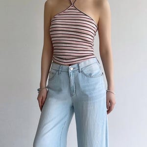 Puede incluir: Un top halter acanalado con rayas horizontales en tonos rosa, blanco y negro, combinado con pantalones vaqueros de mezclilla azul claro. El top tiene un cuello alto con tirantes finos que rodean el cuello, y los vaqueros tienen cierre de bot&oacute;n.