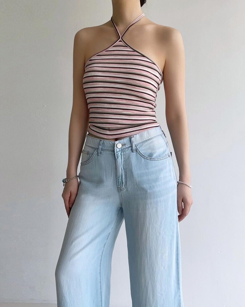 Puede incluir: Un top halter acanalado con rayas horizontales en tonos rosa, blanco y negro, combinado con pantalones vaqueros de mezclilla azul claro. El top tiene un cuello alto con tirantes finos que rodean el cuello, y los vaqueros tienen cierre de bot&oacute;n.