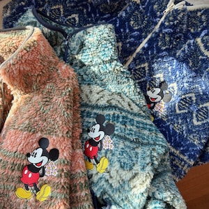 May include: Close-up of three fleece jackets in shades of blue, peach, and teal. Each jacket features a Mickey Mouse patch with a floral accent. The jackets have a zip-up front and a soft, textured appearance.