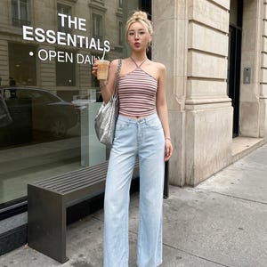 Puede incluir: Un top halter sin mangas con rayas horizontales rosas y blancas combinado con unos vaqueros anchos de color azul claro. El conjunto se complementa con un bolso de hombro plateado metalizado, sandalias transparentes y gafas finas.