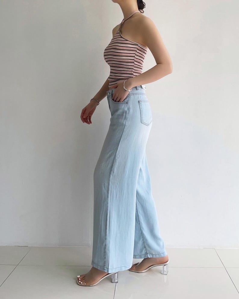 Puede incluir: Un top halter sin mangas con rayas horizontales en tonos rosa, blanco y negro, combinado con pantalones vaqueros de mezclilla azul claro de corte ancho. Los pantalones tienen un dise&ntilde;o cl&aacute;sico de cinco bolsillos con cierre de bot&oacute;n y cremallera.