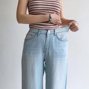 Puede incluir: Vaqueros de mezclilla azul claro con dise&ntilde;o de cintura alta, cierre cl&aacute;sico de bot&oacute;n y cremallera, bolsillos delanteros y corte de pierna recta. Los vaqueros se combinan con una camiseta sin mangas con rayas horizontales rosas, blancas y negras.