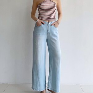 Puede incluir: Un top halter de rayas horizontales en rosa claro y blanco con tirantes oscuros finos, combinado con vaqueros de mezclilla azul claro de pierna ancha con cierre de cremallera, bot&oacute;n y cinco bolsillos, junto a sandalias de tac&oacute;n transparentes.