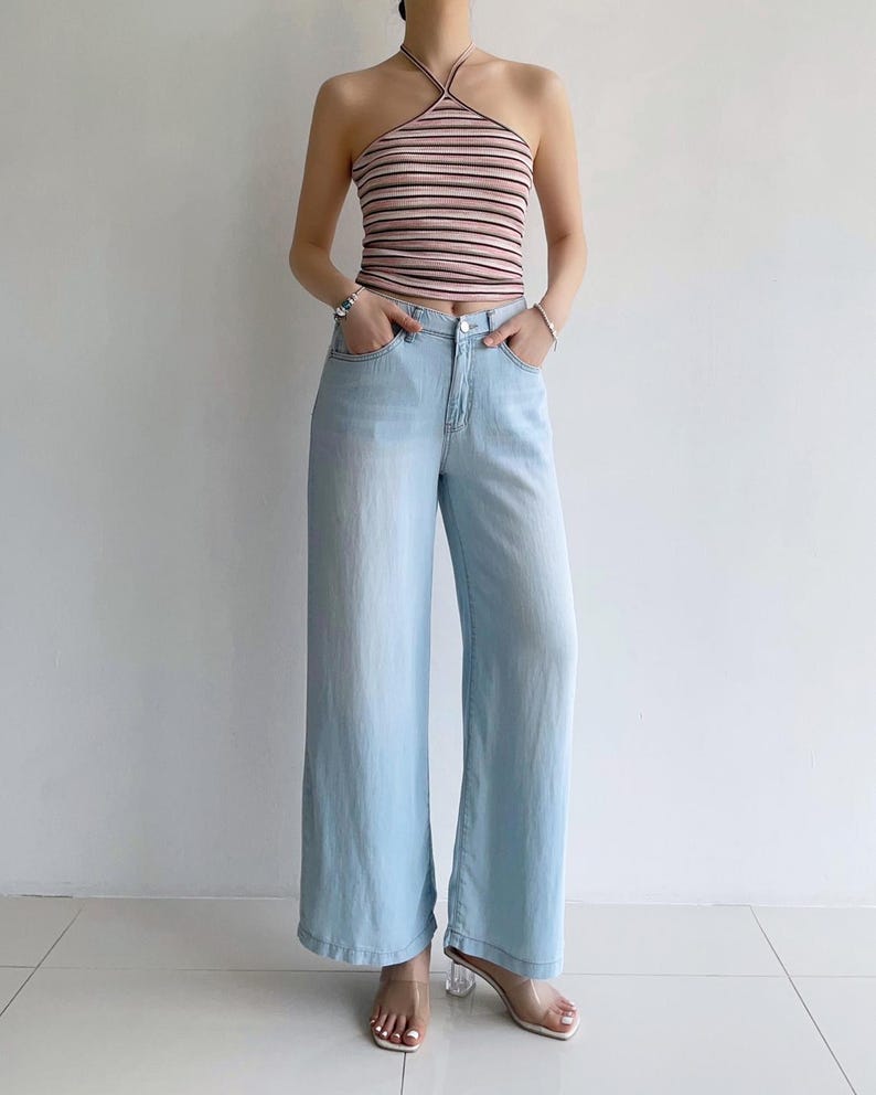Puede incluir: Un top halter de rayas horizontales en rosa claro y blanco con tirantes oscuros finos, combinado con vaqueros de mezclilla azul claro de pierna ancha con cierre de cremallera, bot&oacute;n y cinco bolsillos, junto a sandalias de tac&oacute;n transparentes.