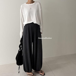 May include: A long-sleeved white crewneck shirt paired with dark charcoal gray wide-leg trousers featuring gathered elastic cuffs at the ankles. A black handbag with a knotted handle is held at the side. The text CocoandEunice is visible.