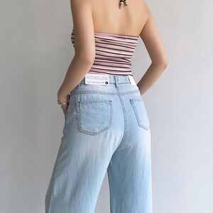 Puede incluir: Un top halter con rayas horizontales en tonos rosa, blanco y negro con cierre de lazo, combinado con vaqueros de mezclilla azul claro. Una etiqueta rectangular blanca con el texto HOWLUK est&aacute; unida a la cintura de los vaqueros.