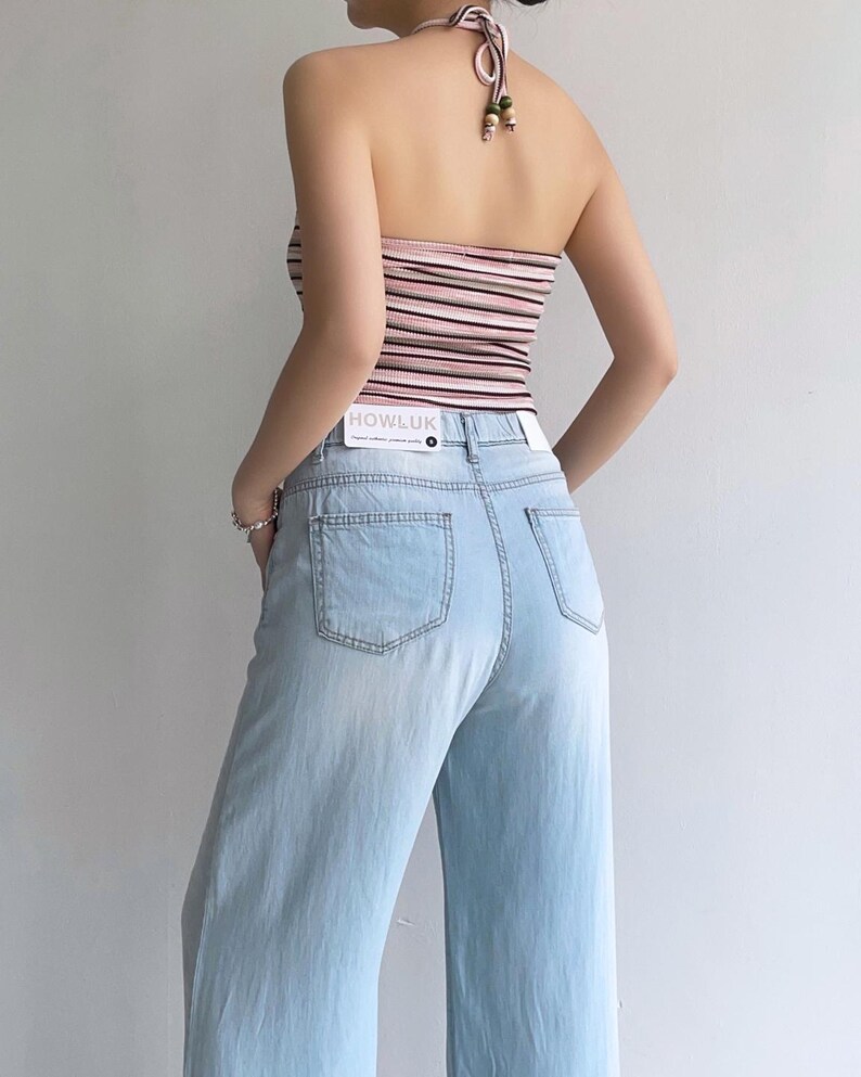 Puede incluir: Un top halter con rayas horizontales en tonos rosa, blanco y negro con cierre de lazo, combinado con vaqueros de mezclilla azul claro. Una etiqueta rectangular blanca con el texto HOWLUK est&aacute; unida a la cintura de los vaqueros.