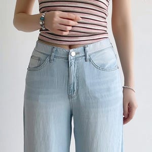 Puede incluir: Un pantal&oacute;n de mezclilla azul claro con dise&ntilde;o de cintura alta, cierre de bot&oacute;n plateado, bolsillos delanteros y cremallera cl&aacute;sica. El pantal&oacute;n se combina con una blusa sin mangas de punto acanalado con rayas horizontales en rosa, blanco y negro.