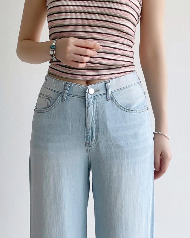 Puede incluir: Un pantal&oacute;n de mezclilla azul claro con dise&ntilde;o de cintura alta, cierre de bot&oacute;n plateado, bolsillos delanteros y cremallera cl&aacute;sica. El pantal&oacute;n se combina con una blusa sin mangas de punto acanalado con rayas horizontales en rosa, blanco y negro.