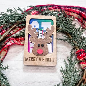 May include: A wooden Christmas card with a reindeer cutout and the text "Merry & Bright". The card is decorated with a red and black plaid ribbon and a green pine branch.