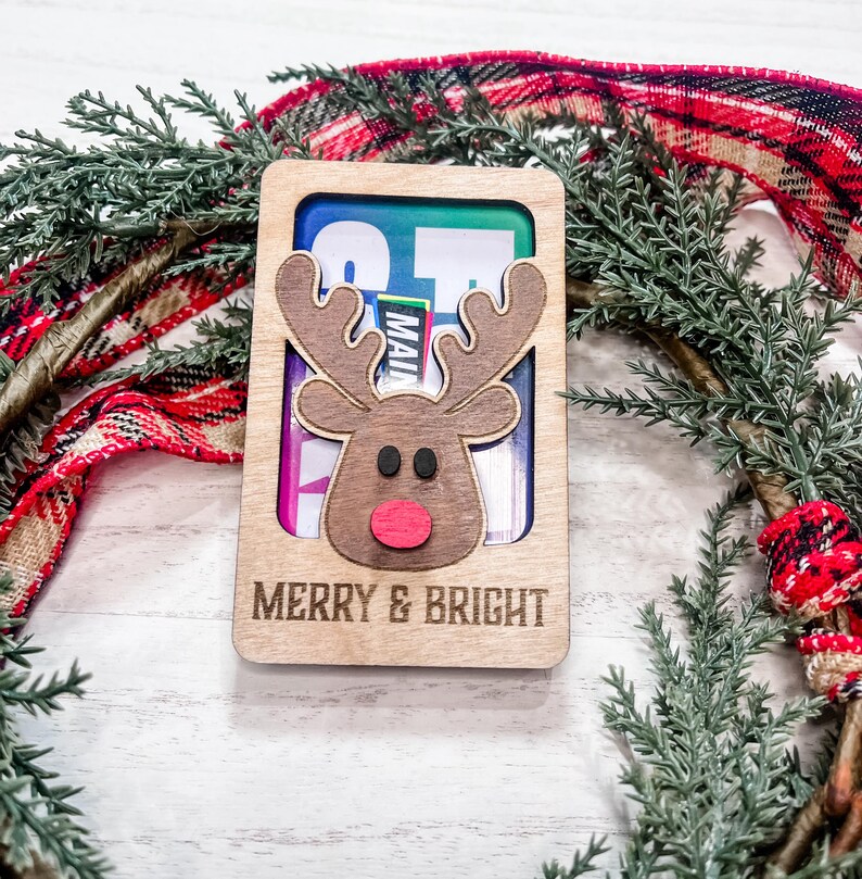 May include: A wooden Christmas card with a reindeer cutout and the text "Merry & Bright". The card is decorated with a red and black plaid ribbon and a green pine branch.
