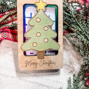 May include: A wooden Christmas ornament with a green Christmas tree cutout and the words "Merry Christmas" engraved on the bottom. The tree has a yellow star on top and white circles for ornaments.