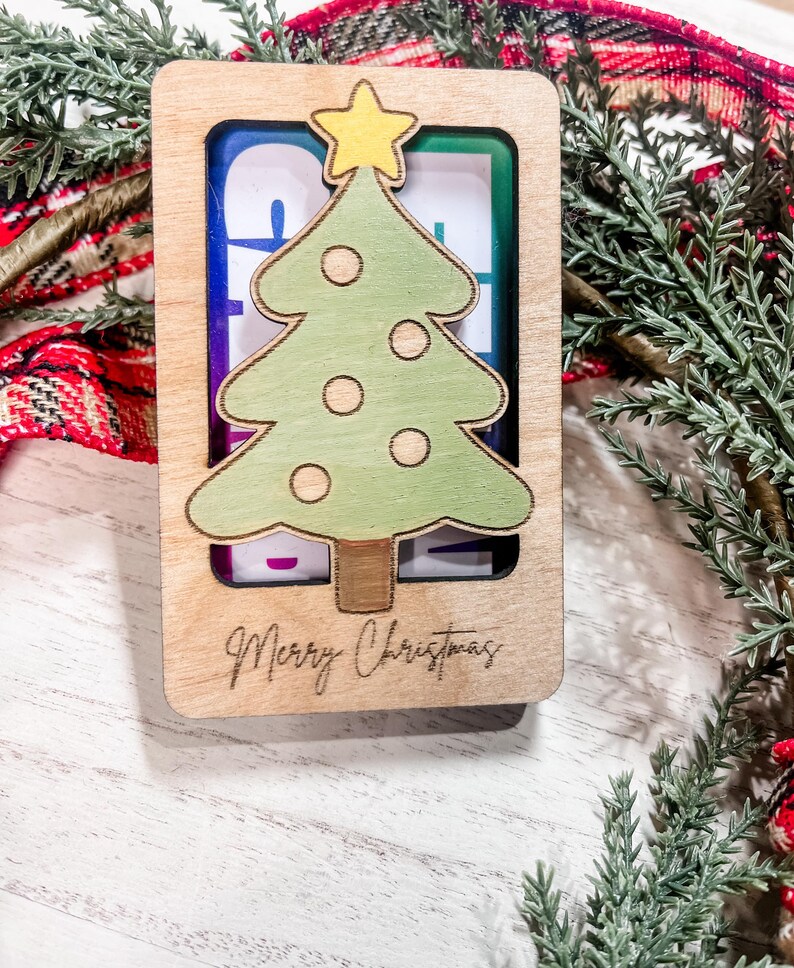 May include: A wooden Christmas ornament with a green Christmas tree cutout and the words "Merry Christmas" engraved on the bottom. The tree has a yellow star on top and white circles for ornaments.