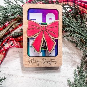 May include: A wooden gift card holder with a red bow and the text "Merry Christmas" engraved on the front. The holder has a clear window to display a gift card.