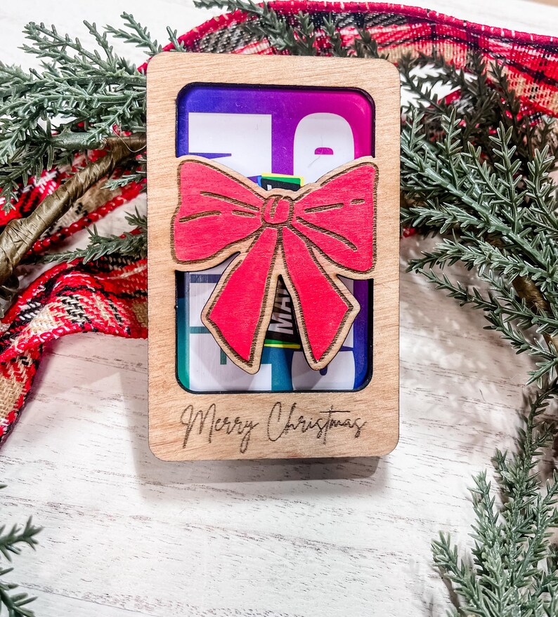 May include: A wooden gift card holder with a red bow and the text "Merry Christmas" engraved on the front. The holder has a clear window to display a gift card.