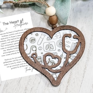 Op de afbeelding: Een houten hartvormig ornament met een stethoscoopontwerp en de woorden "The Heart of Healthcare". Het ornament heeft een witte achtergrond met medische illustraties. Een kaart met tekst over verpleegkundigen staat ernaast.