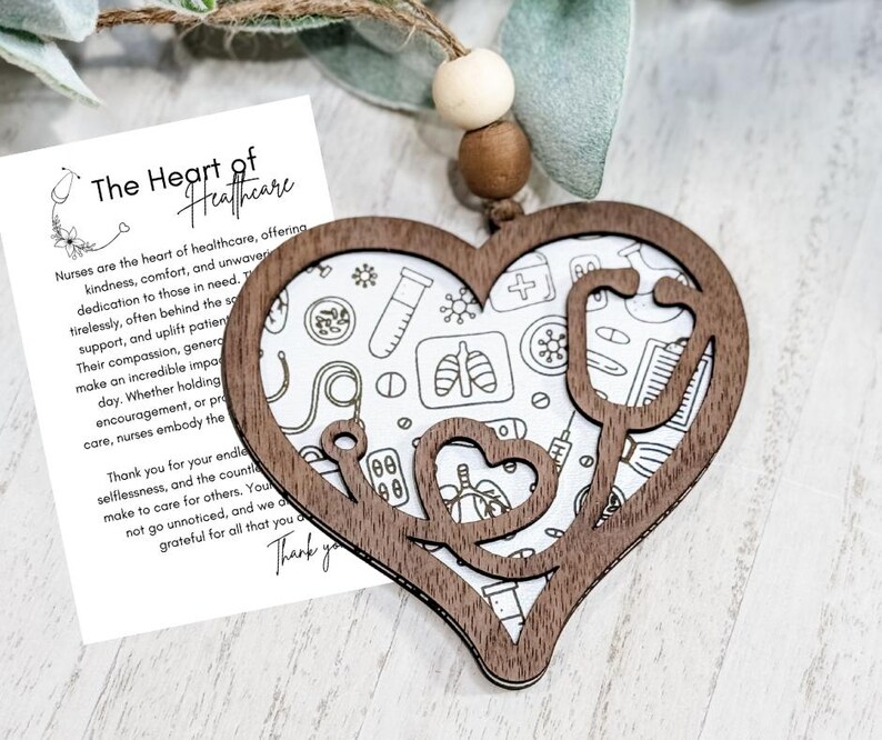 Op de afbeelding: Een houten hartvormig ornament met een stethoscoopontwerp en de woorden "The Heart of Healthcare". Het ornament heeft een witte achtergrond met medische illustraties. Een kaart met tekst over verpleegkundigen staat ernaast.