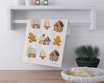 May include: A cream-coloured tea towel featuring a repeating pattern of gingerbread houses, gingerbread men, and Christmas trees. The gingerbread designs are brown and white with red and yellow accents. The towel is hanging on a white shelf.