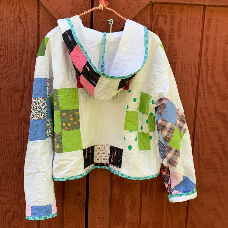 May include: A white hooded jacket with a patchwork design featuring green, blue, pink, and black squares. The jacket has a drawstring hood and a white lining.