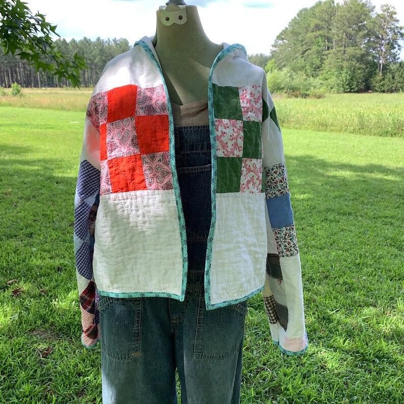 May include: A white quilted jacket with a hood, featuring patchwork panels in red, green, pink, and blue floral patterns. The jacket has a teal trim around the edges.
