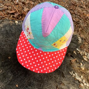 May include: A patchwork baseball cap with a red brim and white polka dots. The crown of the cap is made of different colored fabric scraps, including purple, blue, yellow, and white.