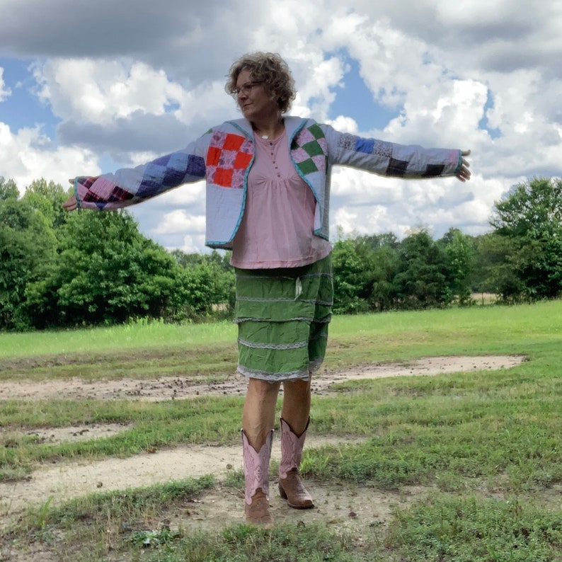 May include: A woman wearing a patchwork jacket with a pink shirt and a green skirt. She is standing in a field with green grass and a cloudy sky.