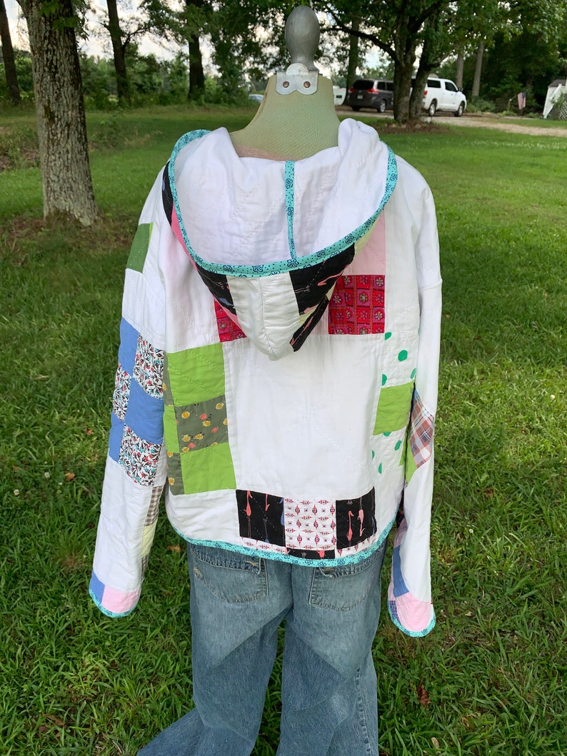 May include: A white quilted jacket with a hood. The jacket is made of different patterned fabrics in shades of blue, green, pink, and black. The jacket has a light blue trim around the hood and a pink trim around the sleeves.