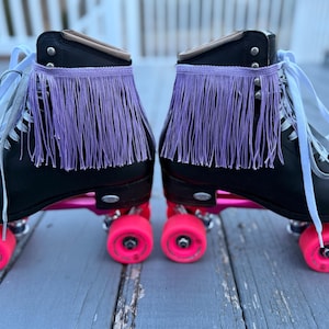 May include: Pair of black roller skates with pink wheels and a purple fringe detail. The skates have white laces and are set against a weathered wooden deck.