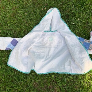May include: A white quilted jacket with a hood. The jacket is made of patchwork fabric with different colored squares. The jacket is lined with a white fabric and has a turquoise trim.